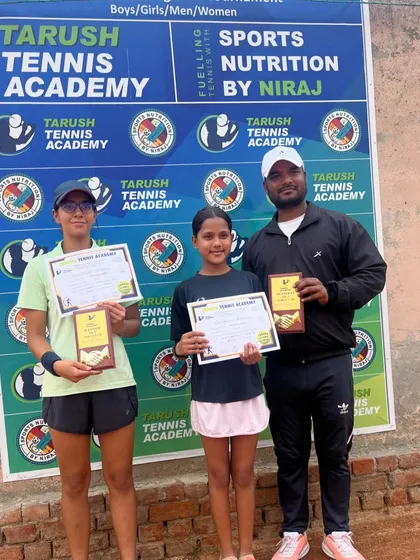 Eshanya Khattar and another player pose with their coach and awards at the Tarush Tennis Academy AITA event, in front of the official tournament backdrop.