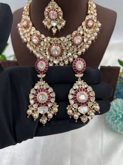 A close-up of the 'Zarista' premium moissanite and kundan earrings, with a matching necklace in the background. The ruby-red stones are encased in delicate pearl work.