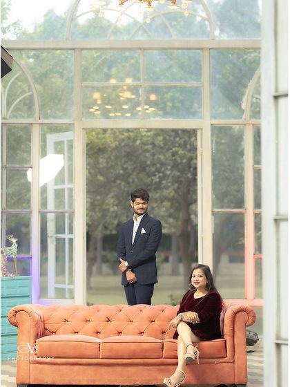 A stylishly composed pre-wedding photo in a glasshouse setting, with the groom standing behind the seated bride, creating a sense of depth and elegance.