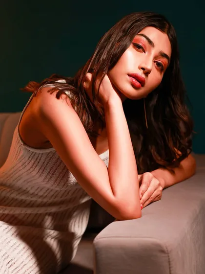 A relaxed portrait of a model in a white top, seated on a couch against a teal wall with warm, soft lighting.