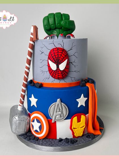 An action-packed two-tiered Avengers cake. This design features Hulk's fist smashing through the top, Spider-Man's mask, Thor's hammer, and other iconic superhero symbols.