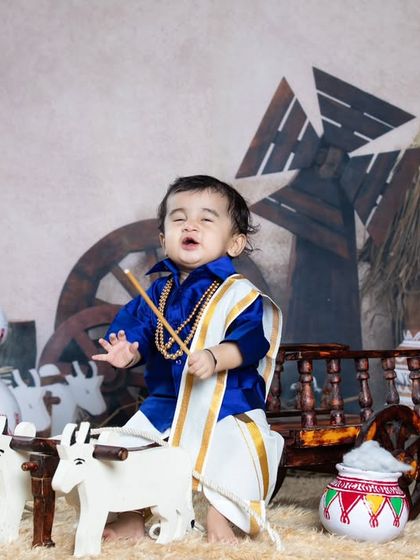 A baby boy dressed in traditional South Indian attire, posing in a rustic village setting.