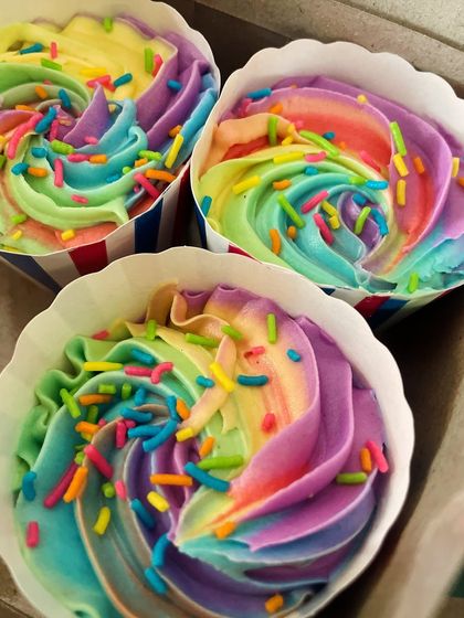 A close-up of three rainbow cupcakes in a box. The vibrant swirls of frosting are perfect for celebrating Pride Month or any happy occasion.