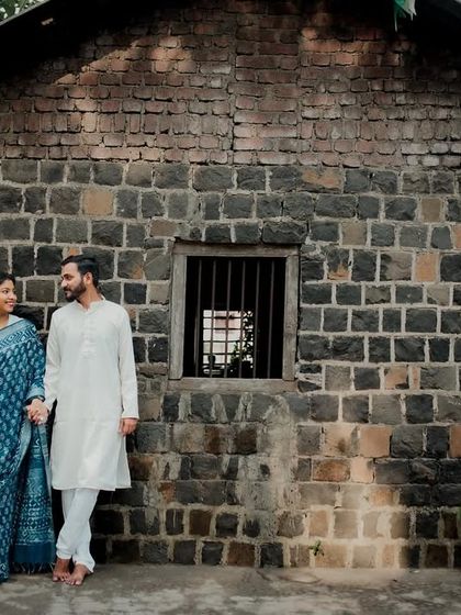 The couple stands together against the backdrop of a rustic stone building, holding hands. This shot combines traditional attire with architectural texture for a unique portrait.