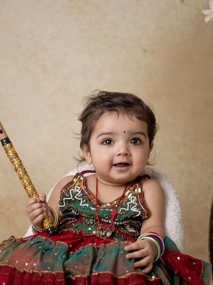 A sweet portrait of a baby girl dressed as Radha, holding her flute and sitting in a mini armchair.