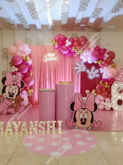 A full-themed Minnie Mouse birthday party decoration for a little one's special day. This setup includes large character cutouts, a pink fabric backdrop, a balloon arch in shades of pink and red, and marquee letters spelling out the birthday girl's name.