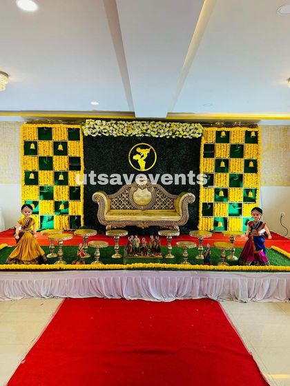 A traditional baby shower stage with a central grass wall panel and marigold grid sides. The setup includes traditional dolls and brass stands for a complete cultural feel.