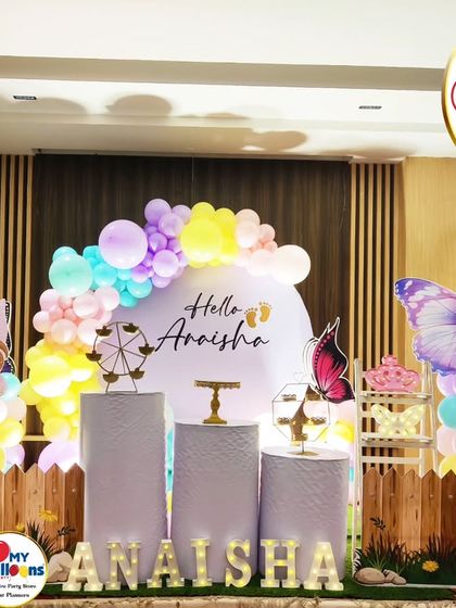 A beautiful fairy and butterfly-themed setup for a baby introduction ceremony. The decor features pastel balloons, a picket fence, and whimsical character cutouts.