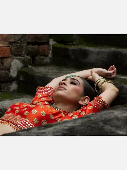 A close-up, artistic shot of a model in a red bridal blouse, lying on stone steps with her eyes closed. This image focuses on the mood and intricate embroidery details of the designer wear.