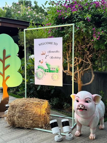 The welcome area for a farm-themed 2nd birthday party. The setup includes a personalized sign, a hay bale, a cute pig statue, and a tree cutout to create a charming barnyard atmosphere.