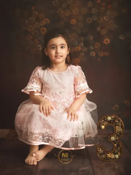 A three-year-old girl in a lovely pink lace dress poses next to a decorative number "3".