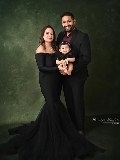 An elegant and formal family portrait. The mother's flowing black gown and the father's sharp suit create a sophisticated look, while their baby girl's smile adds the perfect touch of sweetness.