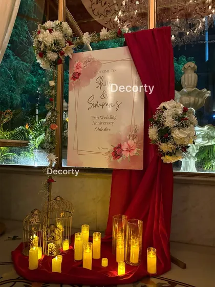 A personalized welcome sign for Shivi and Simran's 15th wedding anniversary. The entrance is elegantly styled with red drapes, candles, and floral bouquets to greet guests warmly.