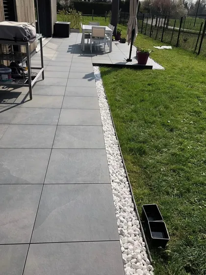 A clean and modern patio design where a border of white pebbles separates the grey tiles from the green lawn.