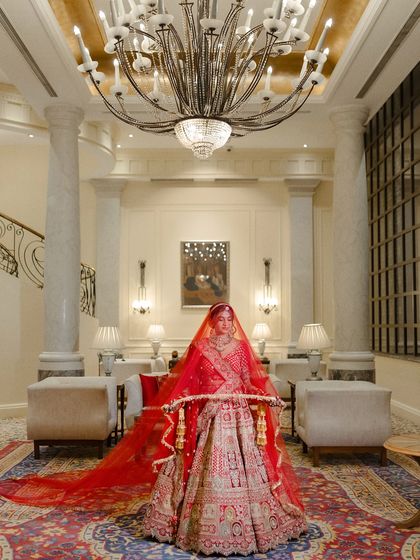 A grand bridal portrait in a luxurious hotel lobby. The bride stands under a massive chandelier, her long red veil creating a dramatic and elegant effect.