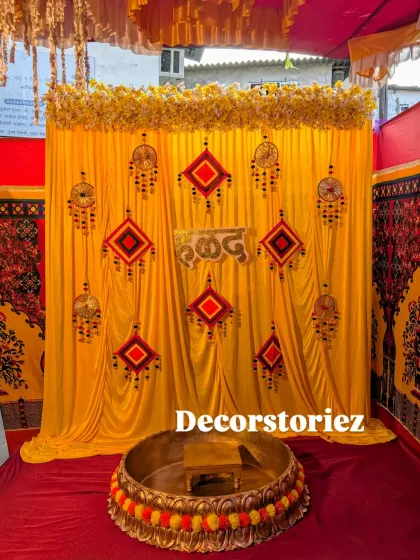 A classic Haldi decoration featuring a bright yellow backdrop, which is a traditional color for the ceremony. The setup is adorned with handcrafted hangings, a 'हळद' sign, and a large brass Urli decorated with marigold flowers.