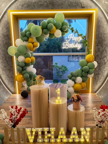 A modern and minimalist setup with an olive green and yellow balloon arch framing a window. A neon sign and marquee letters add a warm glow.