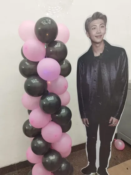 For the ultimate fan, we offer BTS-themed party decorations. This setup includes a life-sized cutout of a band member next to a stylish pink and black balloon pillar.