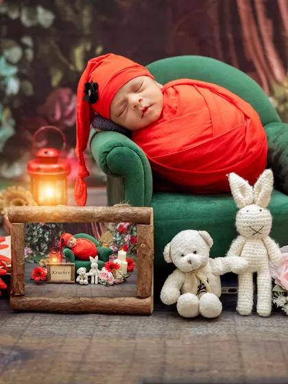 A newborn in a bright red wrap and sleepy hat rests on a miniature green sofa, creating a vibrant and cozy portrait.
