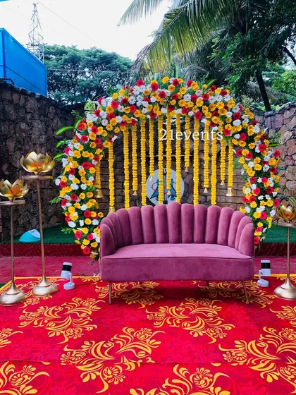 A vibrant and colorful setup perfect for a Mehendi or Haldi ceremony. The backdrop features a floral arch of marigolds and roses with hanging yellow flower strings, set against a modern pink sofa for a fun, festive photo opportunity.