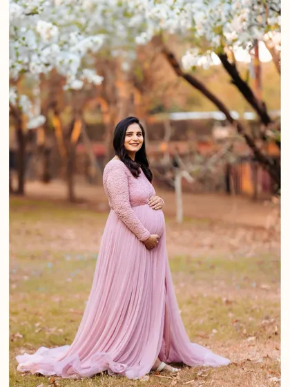 A beautiful solo maternity portrait taken outdoors under blossoming white trees. The mother-to-be glows in a long-sleeved pink gown, her gentle smile and hand on her belly conveying a sense of peaceful joy.