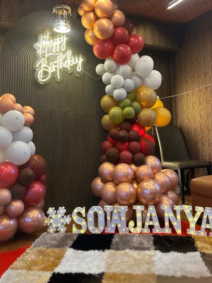 A cozy and stylish birthday corner. The decor features a unique balloon garland in earthy tones against a dark, textured wall, with a 'Happy Birthday' neon sign and marquee letters.