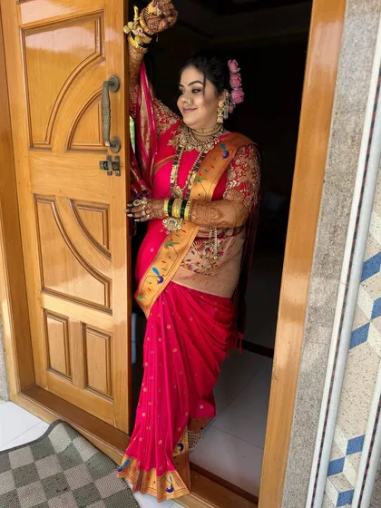 A happy bride in a pink Paithani saree, posing at the entrance of her home.