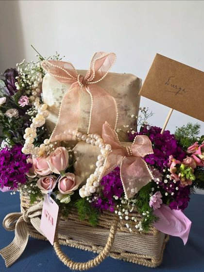 This wicker basket arrangement showcases a classic approach to trousseau gifting. Gifts are wrapped in textured paper and tied with pearl-studded ribbons, surrounded by a mix of fresh pink and purple flowers.