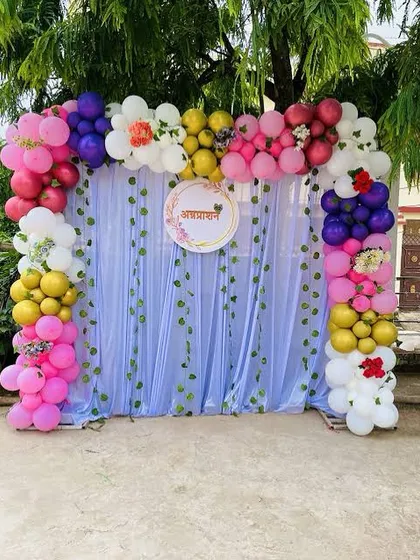 An outdoor backdrop for an Annaprashan ceremony. This setup features a curtain backdrop with hanging vines, a personalized sign, and a vibrant, multi-colored balloon arch.
