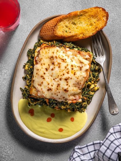 A modern lasagna with layers of spinach and paneer, topped with burnt cheese and served with garlic bread.
