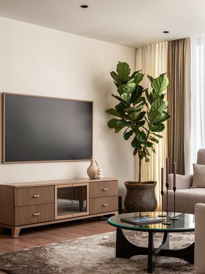 A view of the media unit in the Riviera Maison living room. We designed a custom wooden console with glass-front cabinets to provide storage while maintaining a light and airy feel, complemented by a large fiddle-leaf fig plant.
