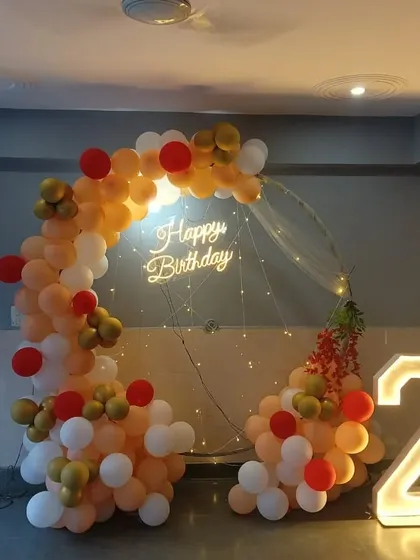 A birthday setup with a semi-circular balloon arch in peach, white, and red. The decoration includes a neon sign, fairy lights, and marquee numbers.