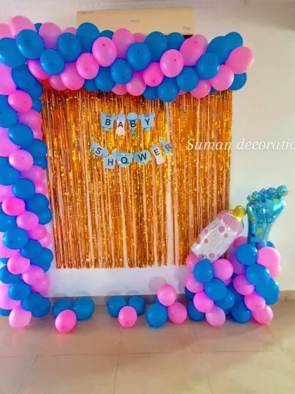 A simple and classic baby shower backdrop. This setup includes a balloon frame in bright pink and blue around a gold tinsel curtain, perfect for a small home celebration.