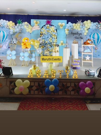A large-scale stage decoration for a baby naming ceremony. The 'Up and Away' theme features a blue and yellow balloon arch, hot air balloon cutouts, and light-up marquee letters for the baby's name.