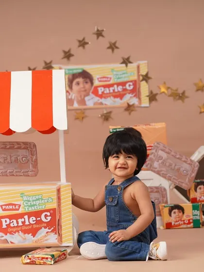 A happy baby girl smiles brightly, sitting beside her Parle-G themed stall in a fun and unique birthday photoshoot.