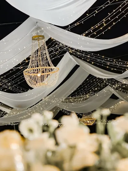 I use lighting and fabric to completely transform a venue. Here, flowing white drapes, strings of fairy lights, and classic crystal chandeliers create a dreamy and romantic ceiling for a reception dinner.