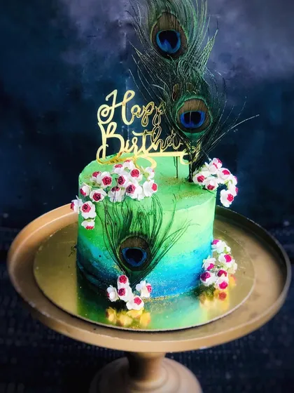 A beautiful Janmashtami cake with a peacock feather theme, featuring vibrant green and blue hues and a golden "Happy Birthday" topper.