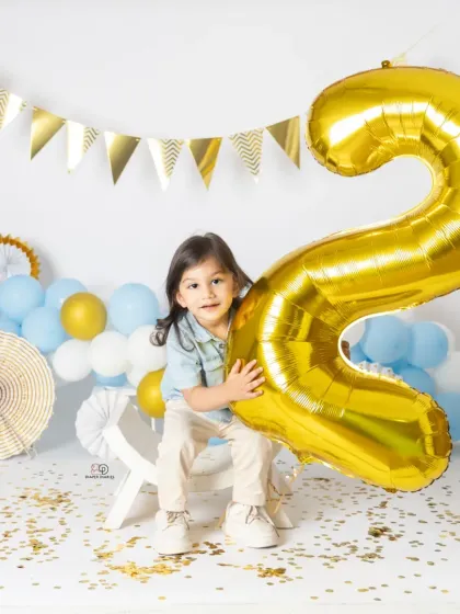 A series of portraits celebrating a second birthday. The toddler poses with a giant gold '2' balloon, surrounded by blue and white balloons and confetti for a festive look.