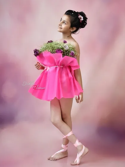 An elegant portrait of a girl in a pink dress made from flowers, holding a bouquet. Her upward gaze and the soft, painterly background give the photo an artistic quality.