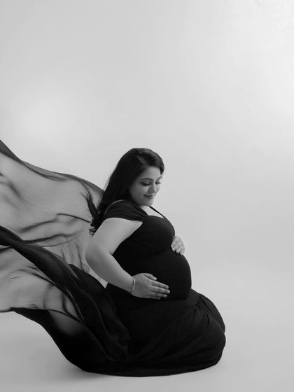 A gentle and introspective black and white portrait. The mother-to-be is seated, looking down at her bump, with the flowing fabric creating a soft, protective circle around her.