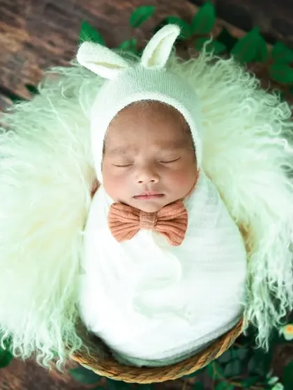 Dressed up and ready for a nap. This baby looks adorable in a white bunny-ear bonnet and a little bow tie, nestled in a fluffy basket.