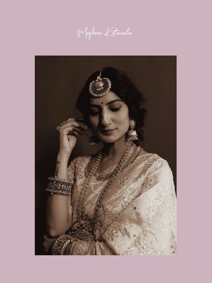 A sepia-toned shot capturing a moment of quiet reflection. The bride is styled in an elegant white saree with intricate jewelry, showcasing a look of timeless grace.
