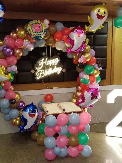 A lively Baby Shark and Cocomelon combination theme. The decoration features a balloon ring with multiple Baby Shark family foil balloons, a Cocomelon balloon, a neon sign, and a light-up number '2'.