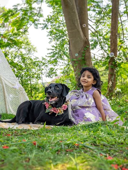 A whimsical outdoor scene with a little girl and her black Lab sitting beside a lace teepee. Bubbles float around them, adding a touch of magic to this beautiful childhood memory being created.