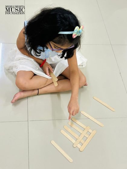 A student uses popsicle sticks to create musical notation on the floor. This hands-on approach makes learning abstract concepts like rhythm and notation tangible and fun.