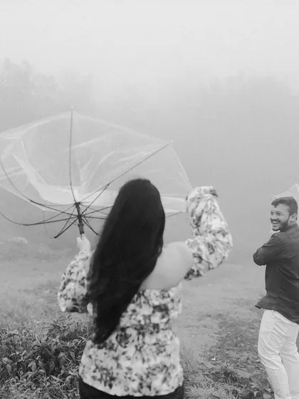 A fun, candid black and white photo from a shoot in Igatpuri, capturing a genuine laugh as the couple plays with an umbrella in the wind and mist.