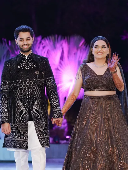 The couple makes their entrance at the sangeet, holding hands and waving. Their coordinated black outfits and the vibrant purple lighting create a festive mood.