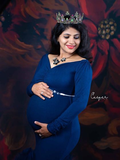 A close-up portrait showcasing a royal blue gown and a beautiful jeweled crown. We focus on capturing the serene expression and radiant glow of the expecting mother against a rich, floral backdrop.