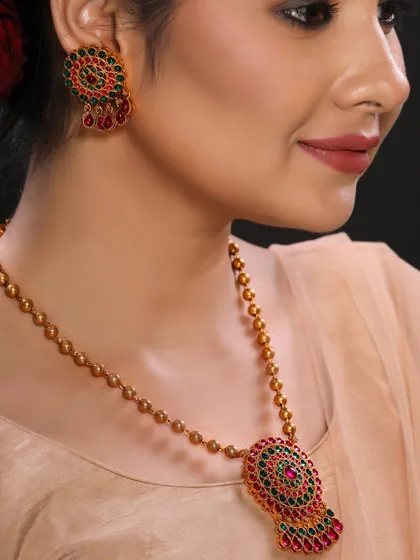 A close-up shot of a model wearing a traditional gold necklace with green and red stones. The soft lighting and elegant pose make this a beautiful image for a jewellery brand's social media or website banner.
