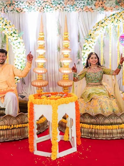 The happy couple at their Haldi ceremony. The backdrop features large floral rings and traditional brass lamps, perfectly complementing the joyous occasion.